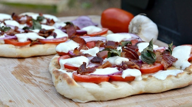 Grilled Bacon and red onion pizza on wooden cutting board with fresh garlic, tomato, and red onion