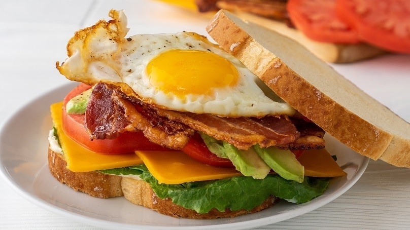 Closeup of a BLT sandwich with egg, avocado and cheese on white plate, fresh tomato slices in background, white table
