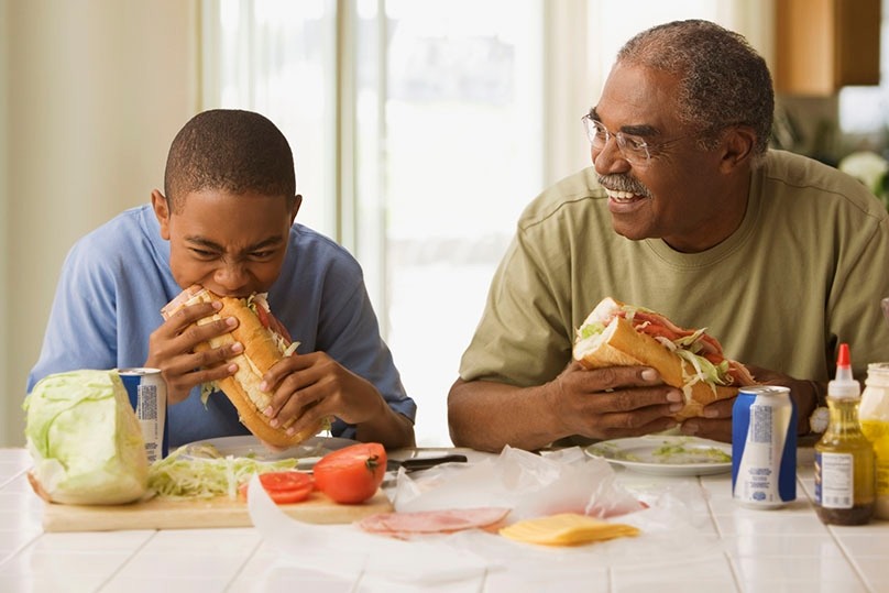 Grandfather and grandson enjoying breakfast sandwiches at the kitchen.counter, white tile counter-top, cutting boards, lettuce, tomato, lunch meat, sliced cheese, condiments, canned drinks