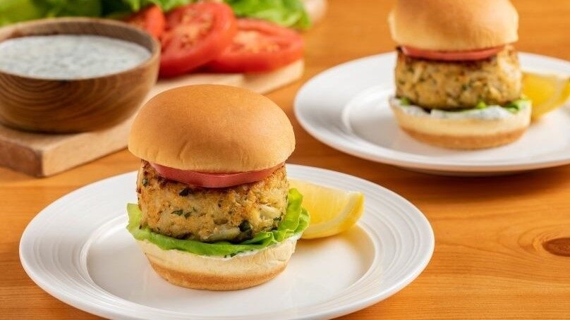 Baked Crab Cake Sandwiches on white plates, wooden table, cutting blard with miscelaneous ingredients