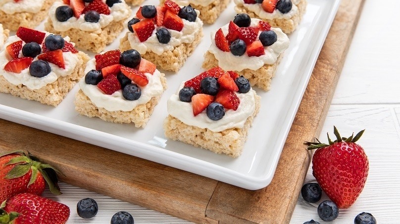 Berry Shortcake Rice Krispies Treats on white serving dish, cuttingboard, strawberries, blueberries, white wood table