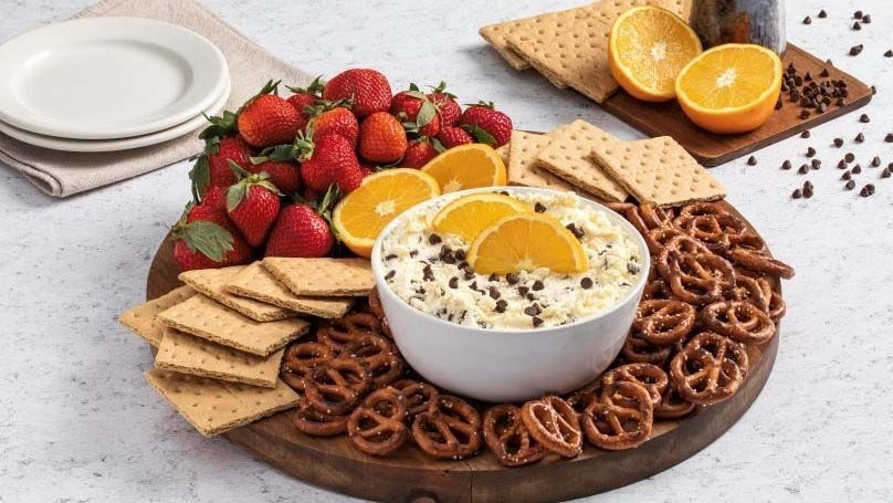 Chocolate Chip Cheesecake Dip in serving bowl surrounded by pretzels, crackers, strawberries, orange slices, chocolate chips, plates, napkin, marble count t