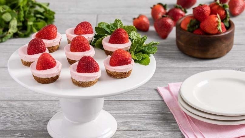 White serving plate with strawberry cheesecake cookie cups and mint leaves, stack of dessert plates, wooden bowl of strawberries, fresh mint leaves, gray wood table