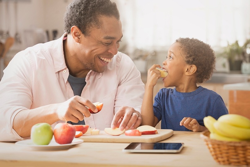 Father and son eating apple slices in kitchen, father smiling at son