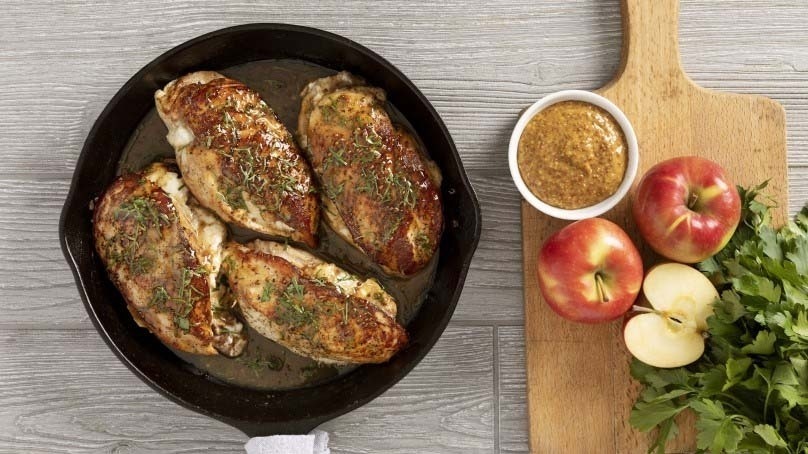 Cast iron pan of apple butter and cream cheese stuffed chicken, cutting board with fresh apples and parsley with serving dish of sauce