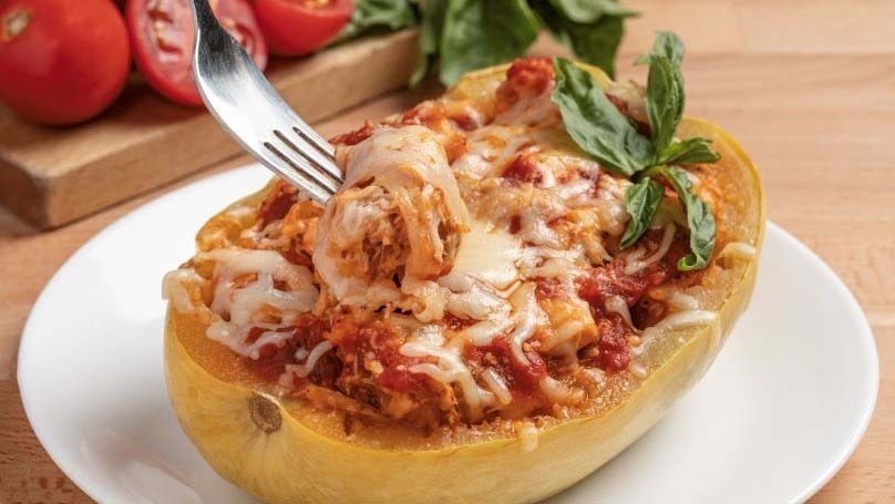 Chicken Parmesan Stuffed Spaghetti Squash on white plate, fresh tomatoes and spinach in background on cutting board, wood table