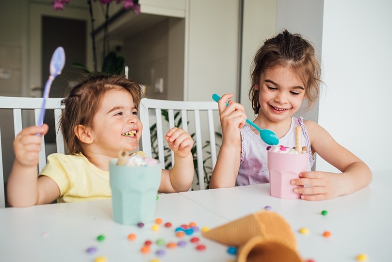 Two young girls laughing enjoying an ice cream treat at white kitchen table