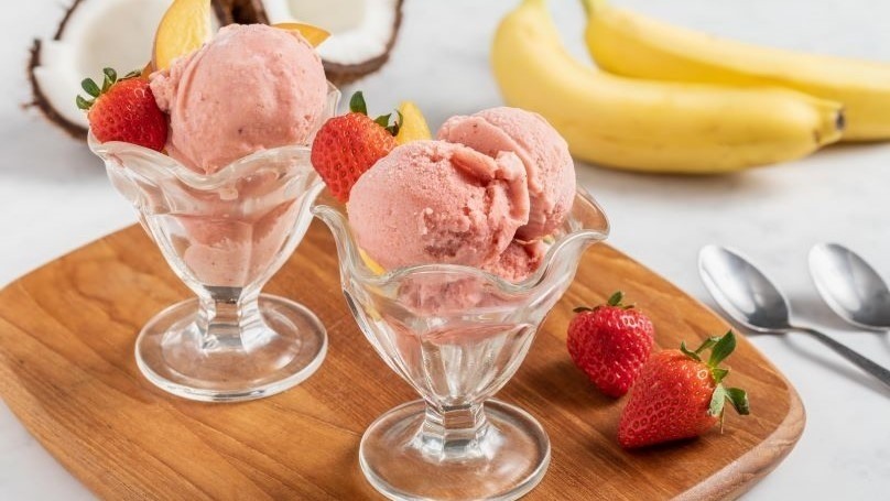 Dairy free strawberry peach ice cream in serving dishes on cutting board with fresh strawberries, bananas and coconuts in background with two spoons