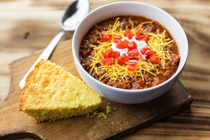 Bowl of Chili, spoon, piece of cornbread, wooden cutting board, wood table