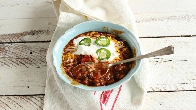Spoon in Bowl of Chili topped with sour cream, cheese, and jalapeno slices, dinner napkin, white wood table