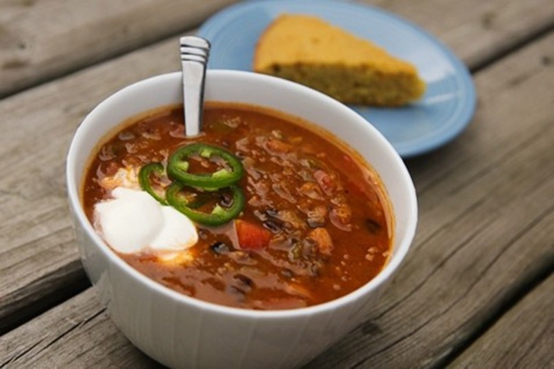 Vegetarian sweet potato chili in white bowl topped with sour cream and jalapeno slices, spoon dipped in chili, slice of cornbread on blue plate, wood table