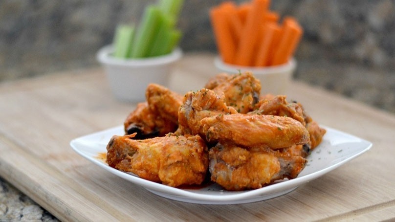 Oven baked wings on white plate, serving bowls of celery and carrot sticks on wooden cutting board, kitchen counter