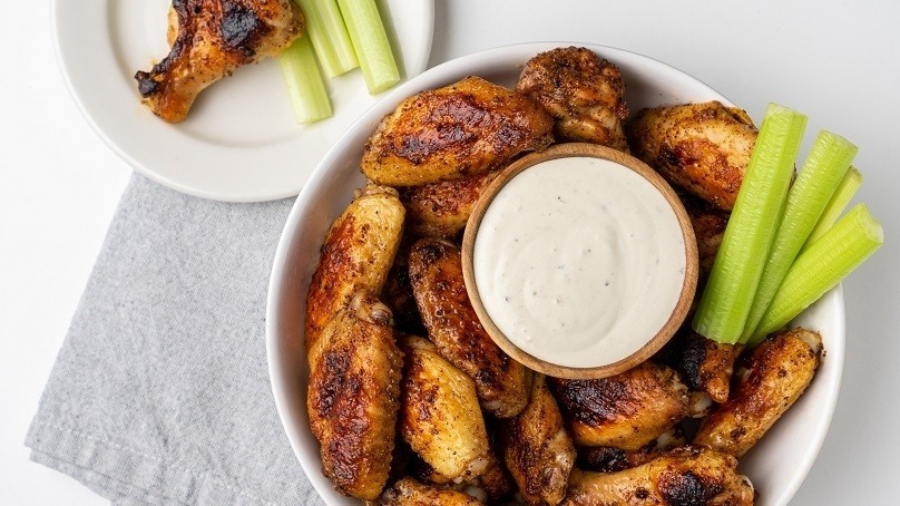 Serving bowl of grilled chicken wings and celery sticks, bowl ranch dressing in the middle, plate of celery sticks and chicken wing, light gray napkin