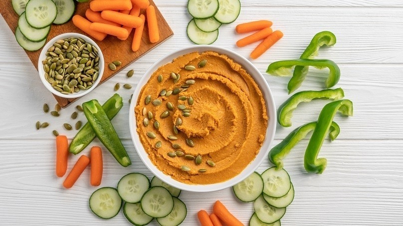 Pumpkin Hummus in bowl surrounded by cut vegetables on white wood table