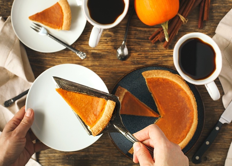 Hand putting a piece of pumpkin pie on white plate, full pie on table below with mugs of coffee, small pumpkin in background, silverware and napkins, wood table background