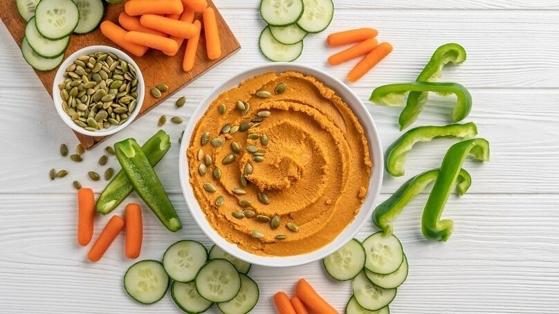 Pumpkin hummus top down view with cut cucumber, carrots, and green peppers, cutting board of fresh cucumbers and carrots, white wood table background