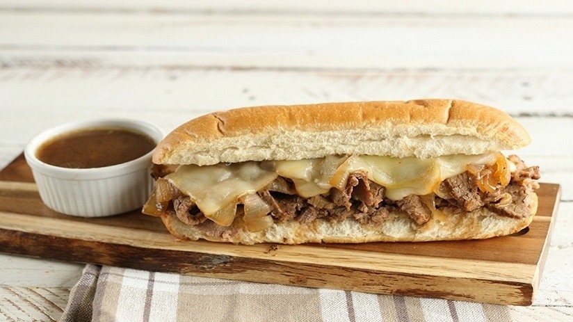 Slow-Cooker French Dip Sandwiche on wood plank with cruet of aus-jus, broiwn checked kitchen towel, rustic wood table