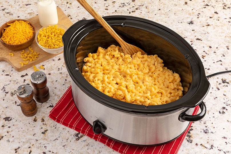 Slow Cooker Mac & Cheese on red towel, kitchen countertop, ingredients on cutting board, salt & pepper, wooden spoon