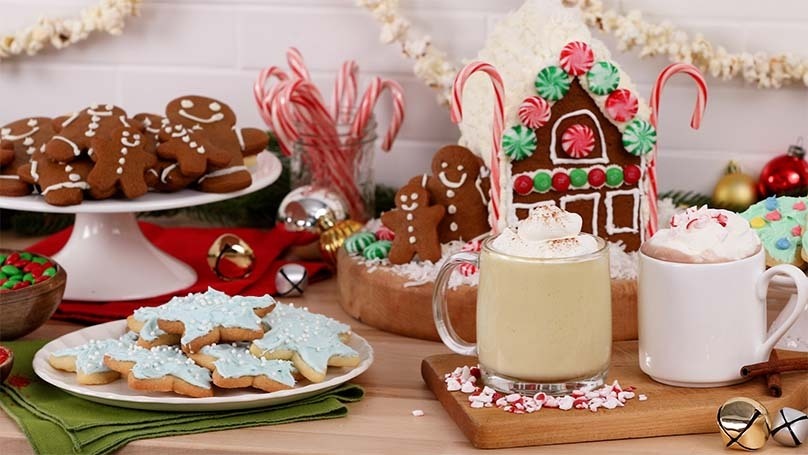 Assortment of holiday desserts on table, decorated gingerbread cookies and house, mugs of hot cocoa with whipped cream, candy canes, etc.