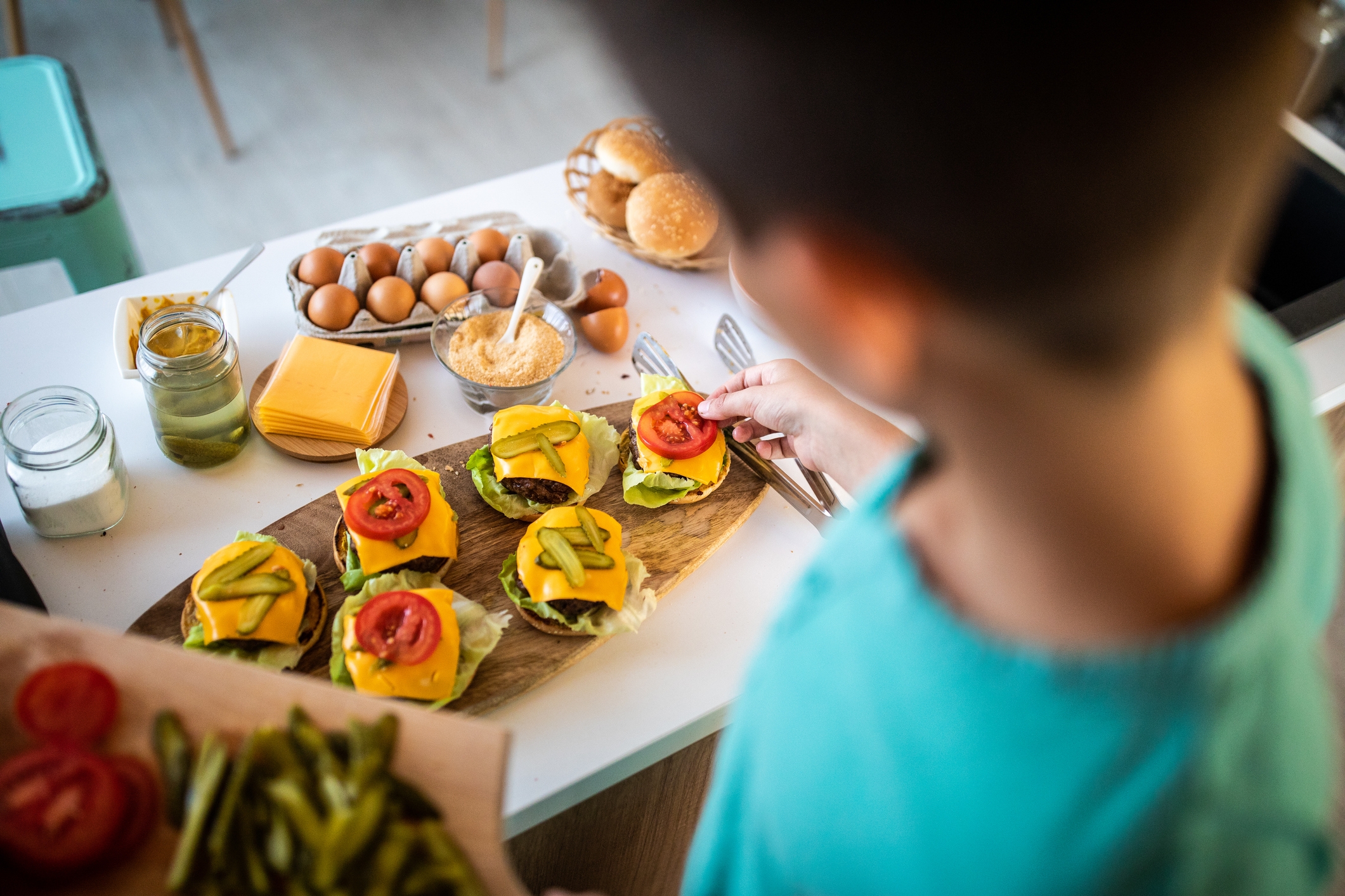 Young boy in kitchen making hamburger sliders with pickles, cheese, tomatoes, and lettuce, on wooden serving dish, white counter, eggs in carton, slices of cheese, jar of pickles, jar of salt, basket of hamburger buns, 