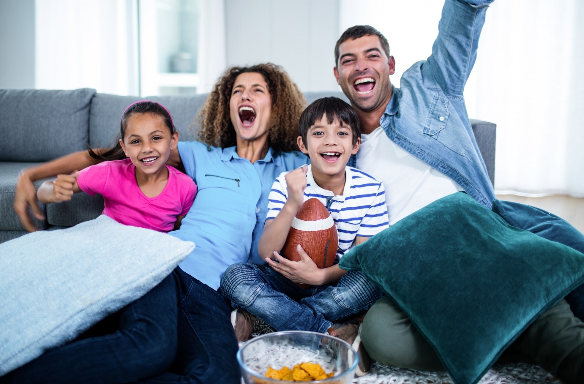 Family on couch watching football game, Mom, Dad, son and daughter celebrating and having fun, son holding football, bowl of chips