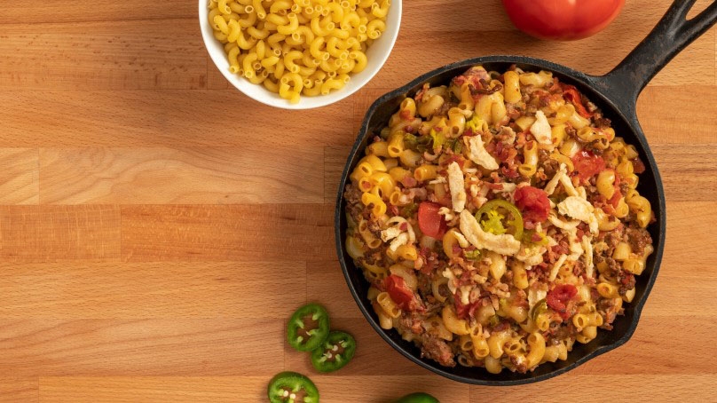 Loaded Mac and Cheese, cast iron skillet, bowl of macaroni noodles, tomato, sliced jalapenos, wood table