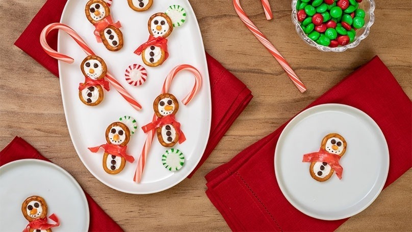 Frosty Snowman Pretzels on serving dish and white olates, bowl of red and green candies, candy canes, red anpkins, wood table