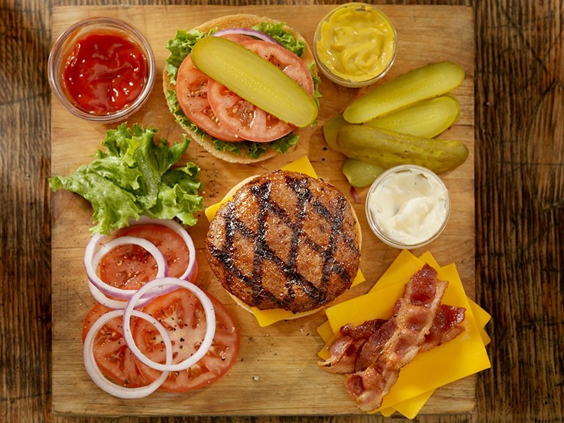Burger ingredients on wood table, tomato slices, sliced pickles, lettuce, bacon strips, cheese slices, mayonnaise, mustard, ketchup