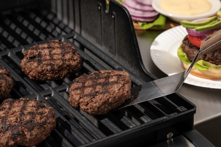 Metal spatula flipping burgers on grill, burger being constructed in background on paper plate