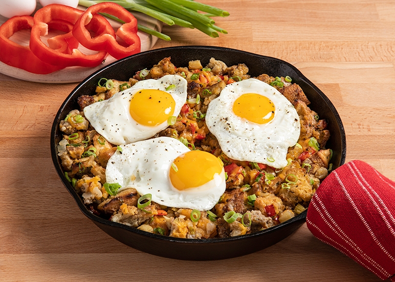 Cast iron pan of leftover stuffing hash with 3 sunny side up eggs on top, fresh red peppers and celery in background