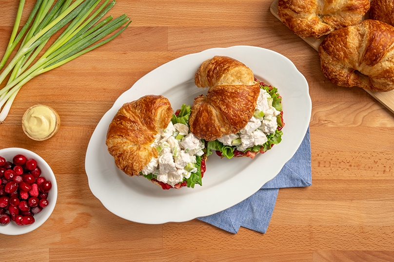 Leftover turkey salad sandwiches on white plate, cranberries, croissants, and fresh celery, wood table background