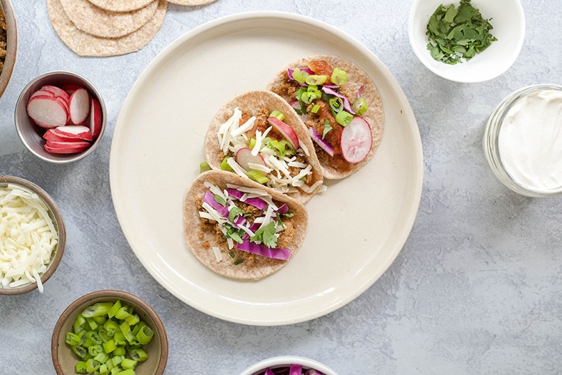 Mini taco bar, 3 mini tacos with turkey, radishes, green peppers, shredded cheese, and red onions on white plate, fresh ingredients in serving dishes surrounding plate