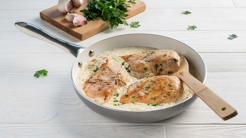 One-Pan Creamy Garlic and Herb Chicken, wooden spoon, cutting board with garloc cloves and parsley, white wood table