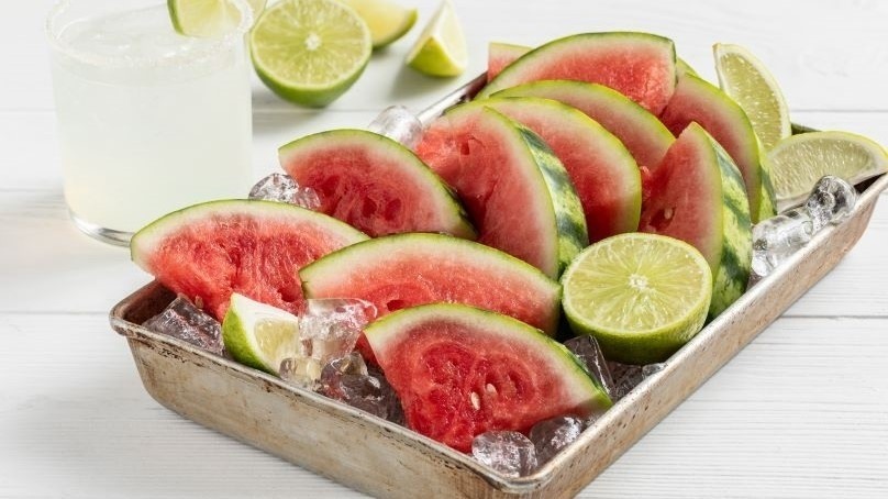 Tray of ice with margarita watermelon slices and fresh halved limes on top, glass of lemonade