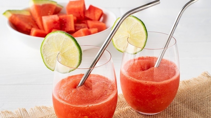 Watermelon Frosé in two glass with lime slice garnish and metal straws, bowl of fresh diced watermelon in background, brown cloth napkin