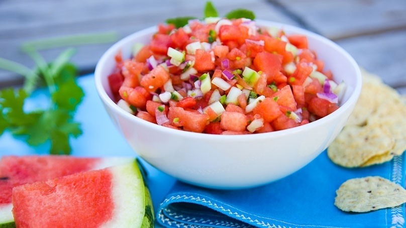 Bowl of watermelon salsa in white bowl, fresh watermelon slices and tortilla chips, blue cloth napkin, wood table