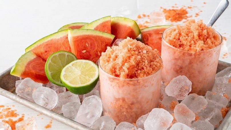 Watermelon lime shaved ice in cups in tray of ice and fresh watermelon slices and halved limes, white table background